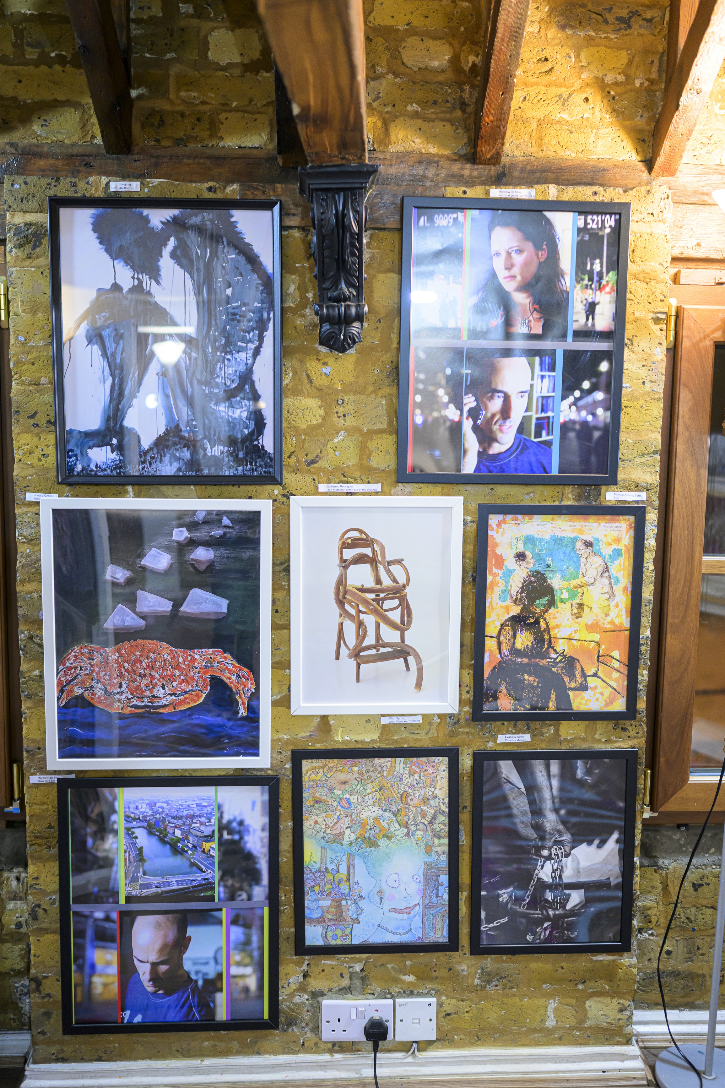 In this image there are eight artworks hanging on a yellowish coloured concrete wall in an art gallery. In the top row, there are two artworks. The one on the left is a figurative oil painting of a very slim body of a human, probably an old man, sitting on a rock with his knees bent and close to his belly. His head falling towards his knees. The painting has blue tones and is characterised by spikes around the man. The second one is a collage created by Matthew da Silva, comprising four six photographs aligned in three by two table. The centre column depicts a woman on top gazing at something or someone, and the bottom one depicts a man talking to someone on his cellphone. These two photographs are taken from a TV show. The middle row shows three artworks, the first is an oil painting of a big sweet looking crab. On its back, rocks of various shape are depicted with a perspective and extends beyond the frame of the painting. The middle painting in the second row is a painting of a wooden chair tangled by its own sections or a long curving piece of wood.